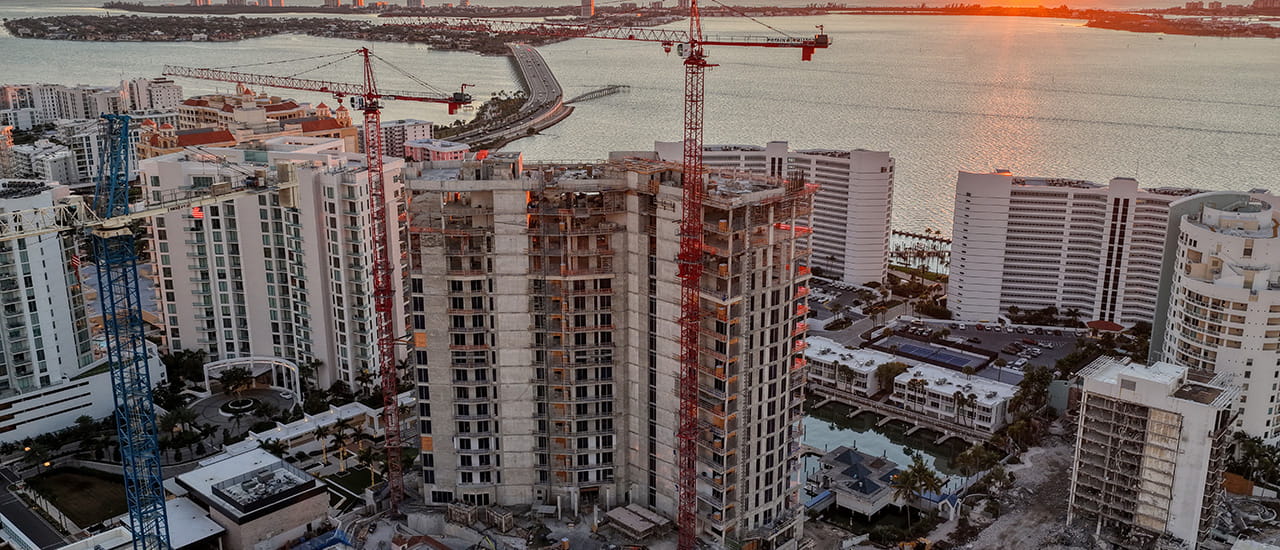 High-rise building under construction with cranes near water at sunset.