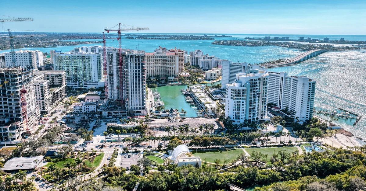 Aerial photo of the Sarasota bayfront skyline with multiple high-rise buildings, construction cranes, a marina, and turquoise bay water under a clear blue sky.