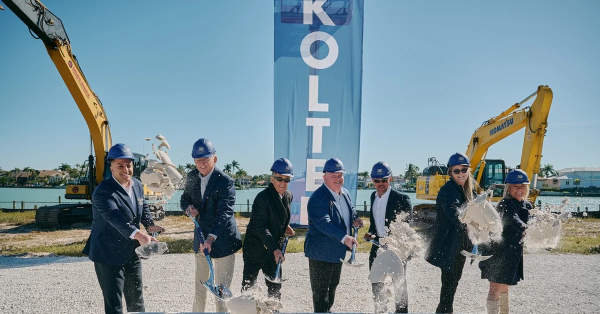 Seven people in business attire and blue hard hats tossing sand with ceremonial shovels at a groundbreaking event, standing behind a sign reading '3300 Gulf Shore Naples,' with excavators and a waterway in the background.