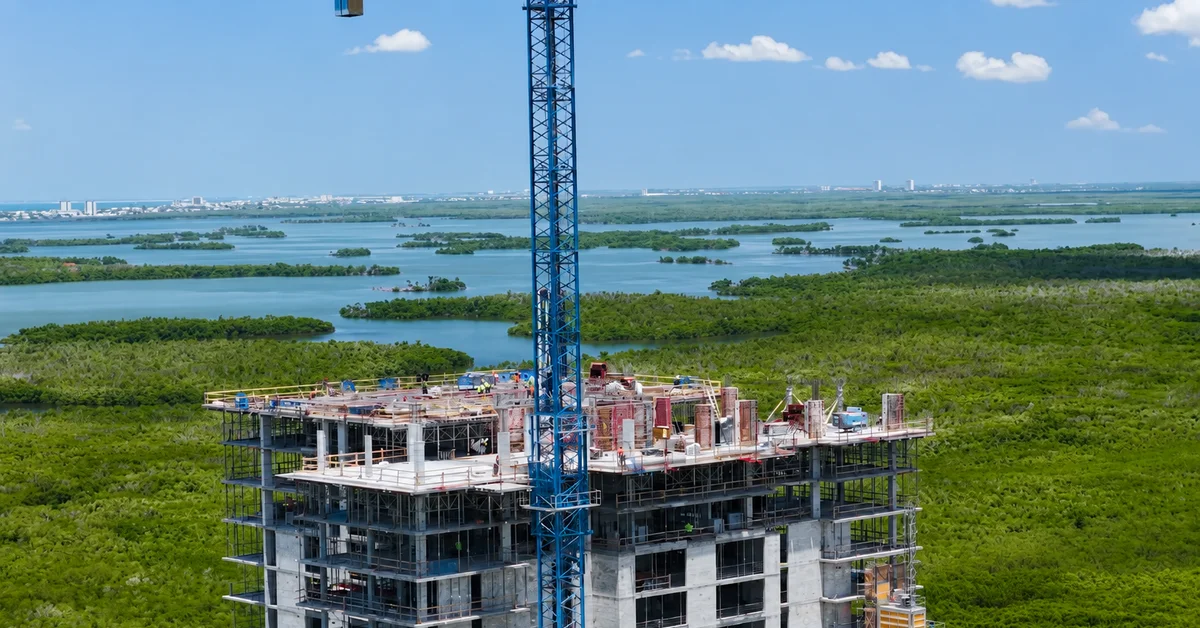 Aerial photo of a concrete high-rise tower under construction with a blue tower crane on top, surrounded by green mangrove islands and blue water, with a coastline visible in the far distance.