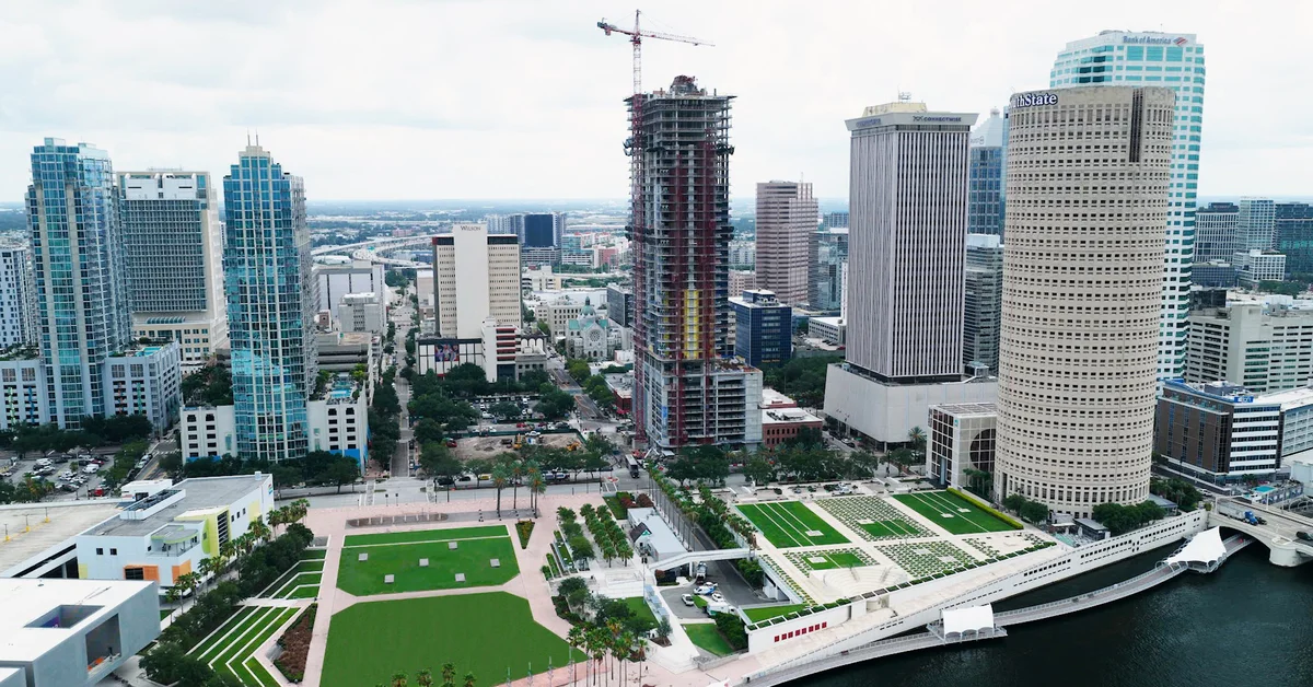 Aerial photo of a tall high-rise tower under construction in downtown Tampa, with a construction crane on top, surrounded by other skyscrapers, a riverfront park, and the Hillsborough River.