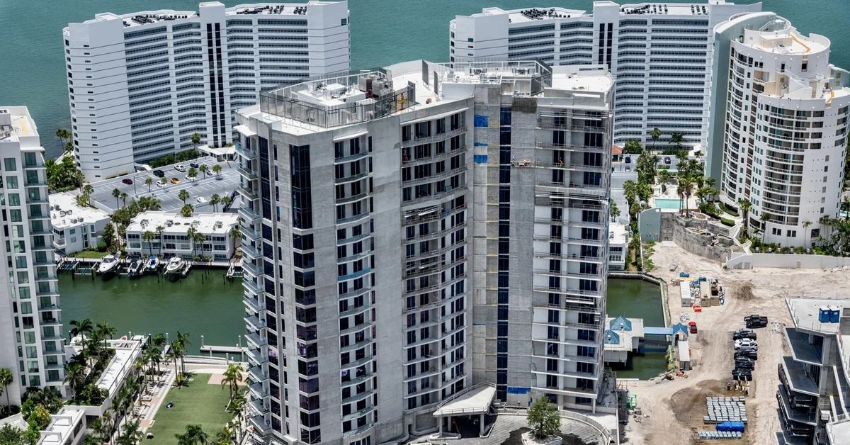 Aerial photo of a high-rise concrete tower under construction on Sarasota Bay, surrounded by white condominium buildings, a marina with boats, and turquoise water.